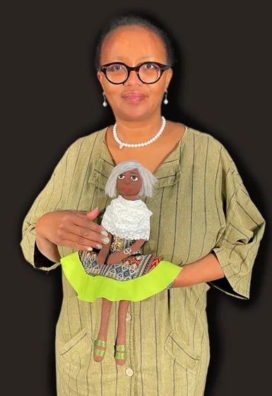 A woman with black hair and perl jewellery is holding a puppet in her hands.