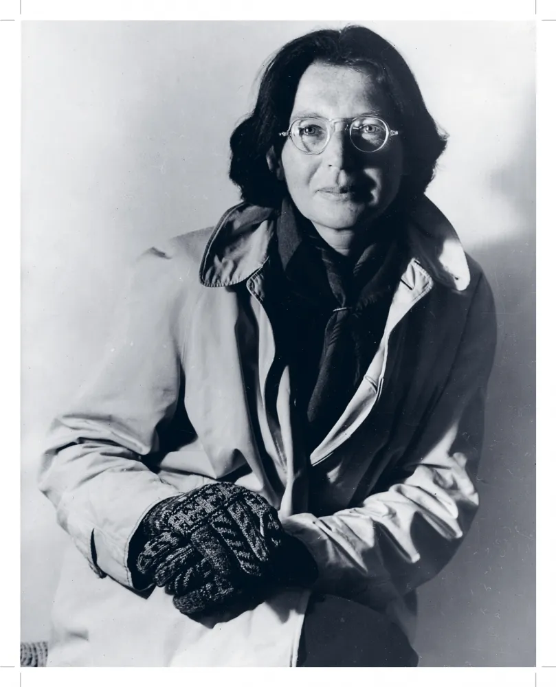Schwarz-weiß-Fotografie einer Frau, die einen hellen Mantel und schwarze Handschuhe trägt und sich damit auf ihrem aufgestellten Knie abstützt.