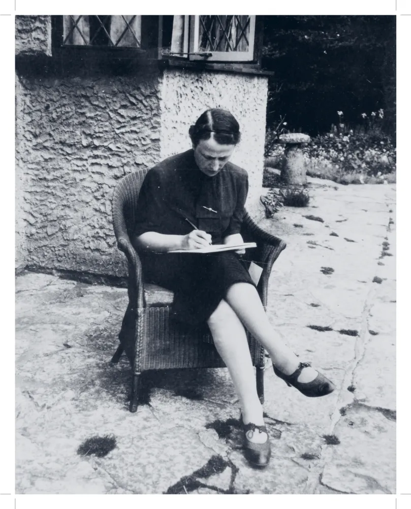 Schwarz-weiß-Fotografie einer Frau, die in einem Korbsessel auf einer Terrasse sitzt, mit einem Stift in der Hand und einem Klemmbrett auf den Knien. Sie blickt auf das Brett.