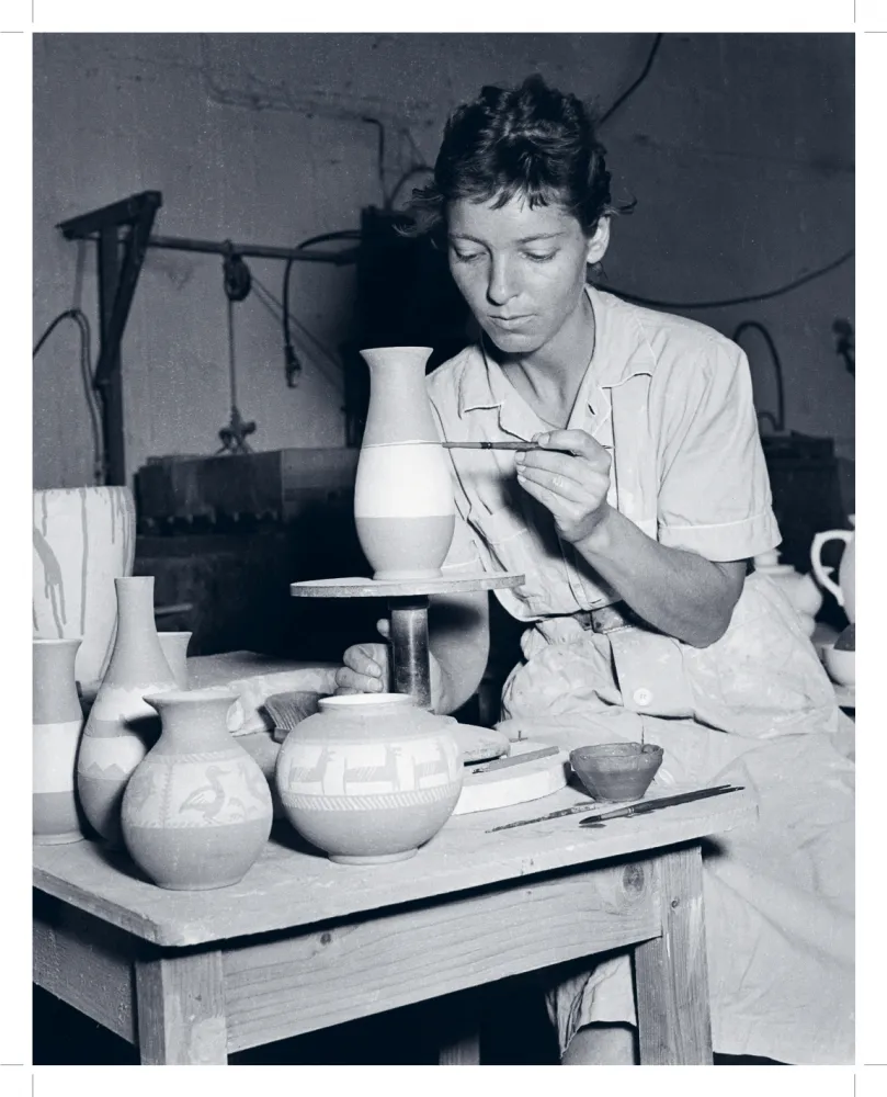 Eine schwarz-weiß-Fotografie zeigt eine junge Frau an einem Tisch mit Keramikgut sitzend. Sie bemalt eine Form, die vor ihr auf einem kleinen Podest steht.
