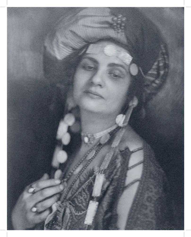 Die sepaifarbene Fotografie zeigt eine Frau mit Turban und einer Tunika und auffälligem Schmuck. Ihr Oberkörper und Gesicht sind zur Kamera gewendet aber ihr Blick geht Richtung Boden.