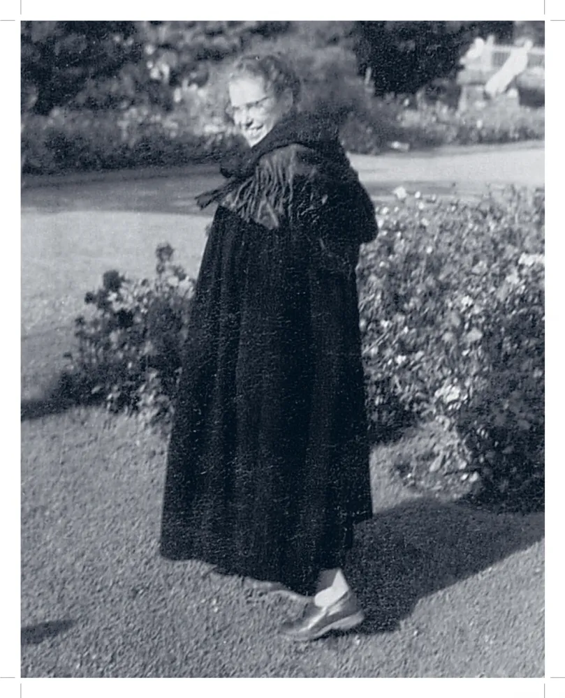 Schwarz-Weiß-Foto einer Frau in einem Garten. Sie trägt einen Mantel, eine Brille und lächselt.