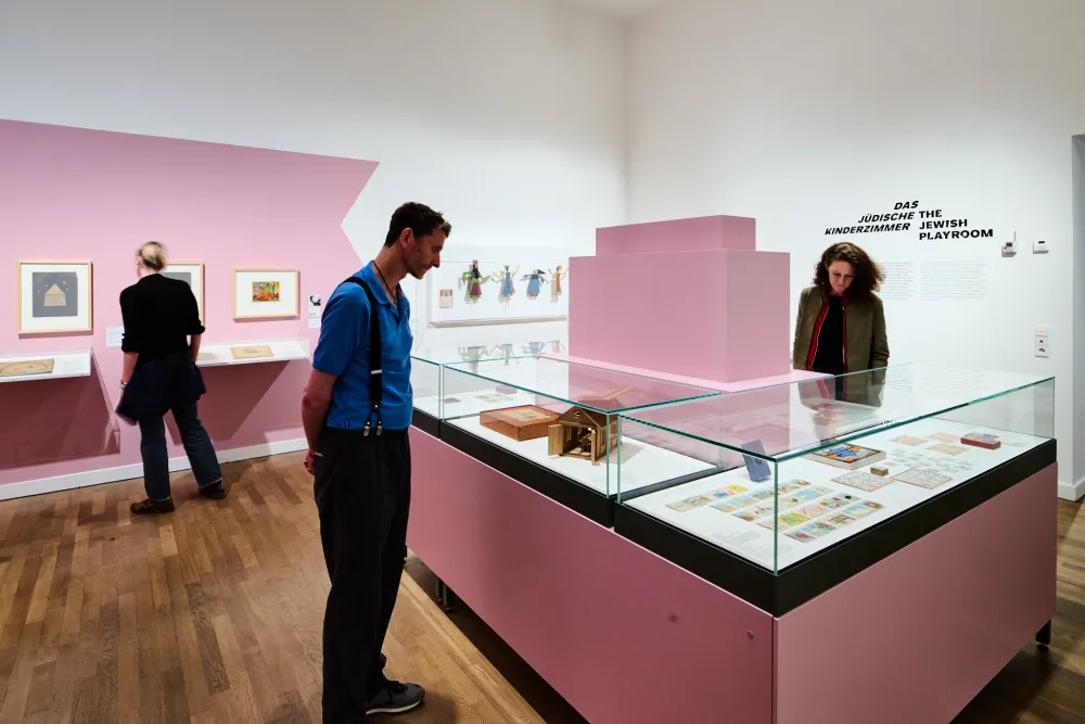 Ausstellungsansicht mit drei Personen und Objekten, die in Vitrinen liegen oder an der Wand hängen.