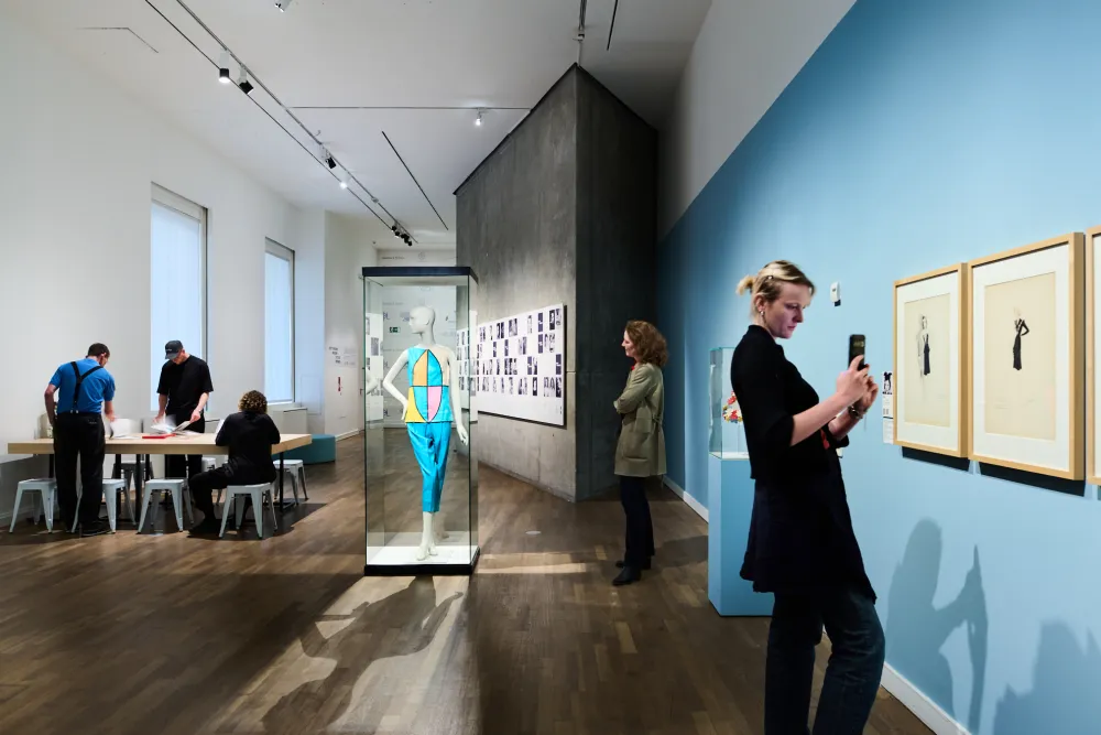 Ausstellungsansicht mit fünf Personen und Objekten, die in Vitrinen liegen und an der Wand hängen. Menschen sitzen und stehen an einem Tisch.