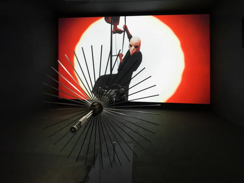 Futuristischer Metallgegenstand, der aussieht wie ein Ventilator oder Satellit, zeigt auf eine gro&szlig;e Filmleinwand. Auf der Leinwand steigen Personen eine Leiter empor. Eine Person hat eine Vogelkopfmaske auf. 