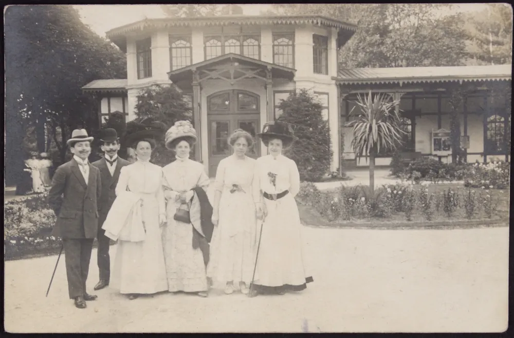 Schwarz-Weiß Bild. Zwei Männer in dunklen Anzügen stehen links. Rechts daneben stehen vier Frauen in hellen Kleidern.