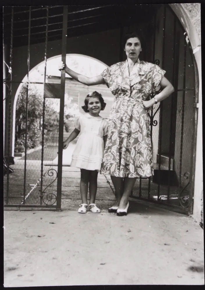 Schwarz-Weiß Bild. Eine Frau mittleren Alter und ein kleines Mädchen stehen neben einem Gittertor vor einem Hauseingang