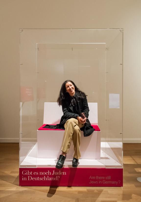 Sharon Adler sitting on a bench in a plexiglass box which is open at the front, on the pedestal is written: Are there still Jews in Germany?