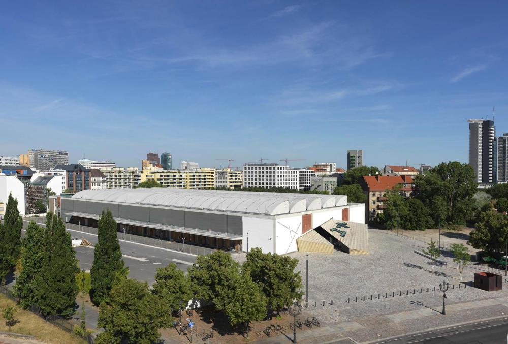 Vogelperspektive auf eine lange, flache Halle, umgeben von Asphaltflächen und Bäumen