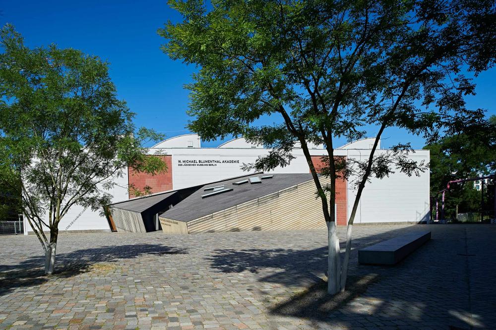 White painted front of the hall with wooden entrance cube and two green trees on the paved forecourt