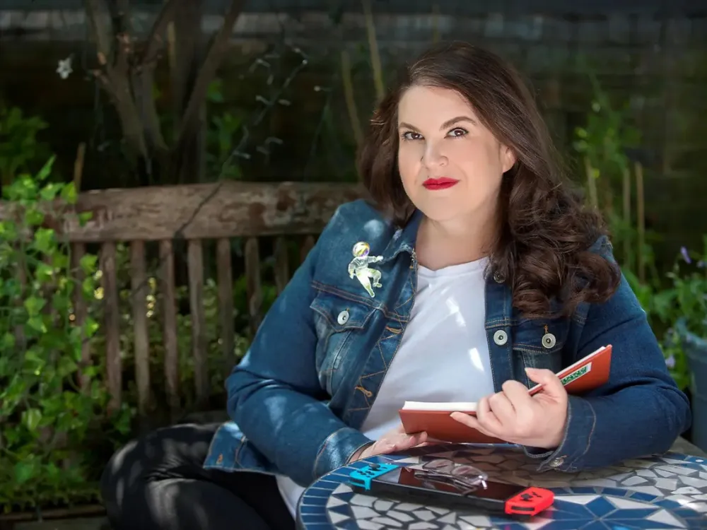 A woman is sitting on a bench with a book in her hand, smiling at the camera.