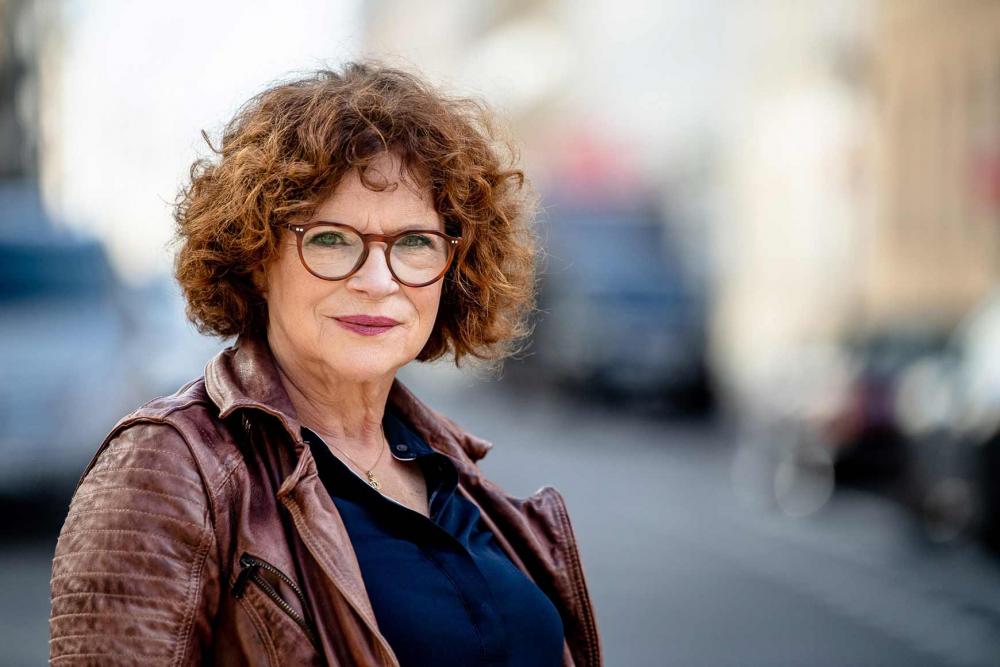 Curly woman with glasses and leather jacket looks at camera.