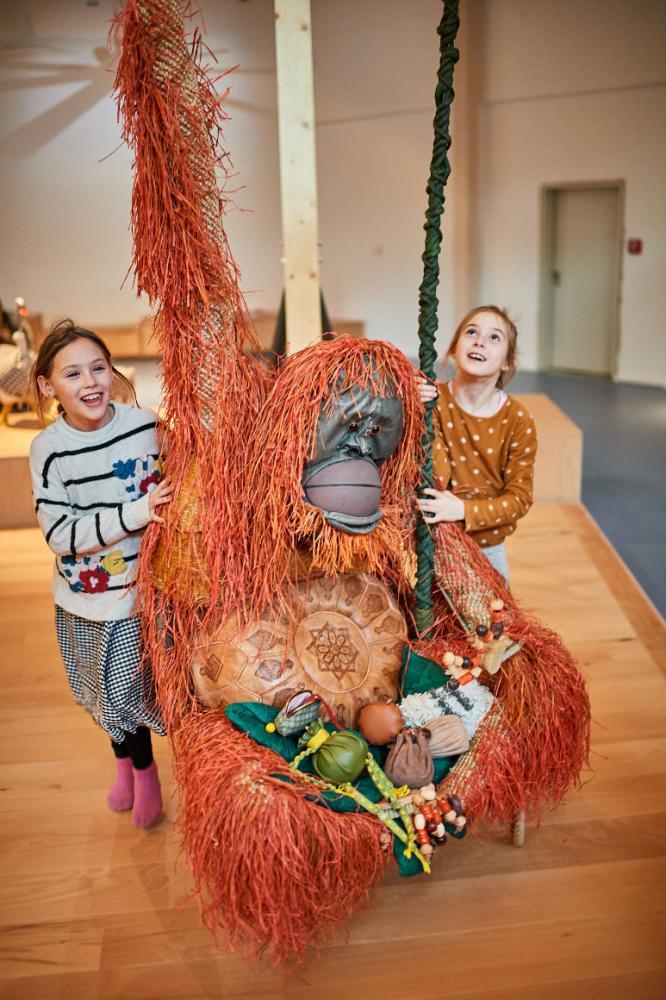 Zwei Kinder stehen neben einem aus Alltagsgegenständen gebauten Orang-Utan, der an der Decke hängt