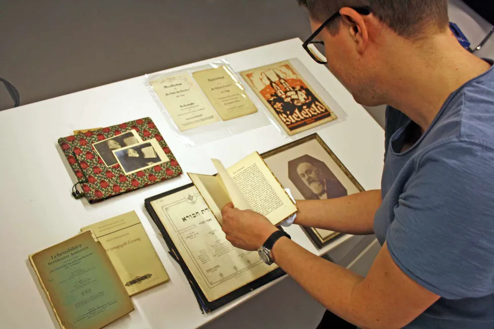 A man is reading an old little book; there are other books and documents on the table.