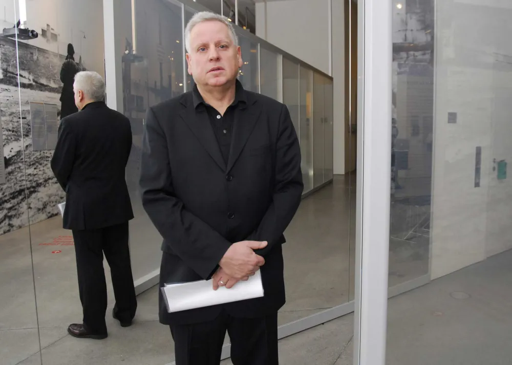 Stehender Mann in schwarzem Anzug vor einer verspiegelten Glasfläche, seine Körperrückseite ist als Spiegelbild zu sehen.