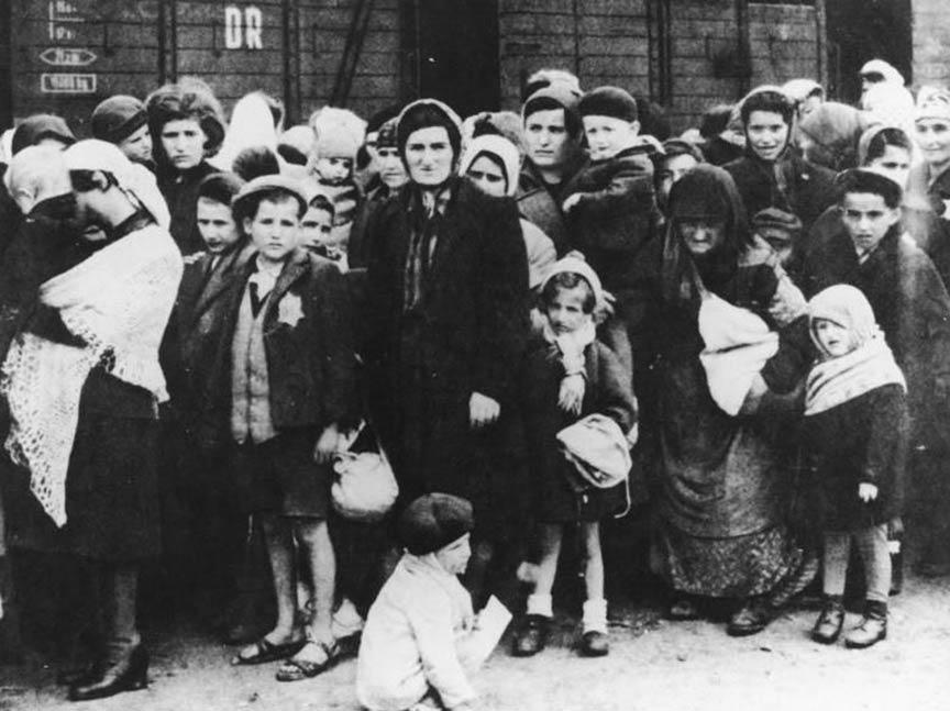 Black and white photograph with numerous people of different ages, wearing the Jewish star and standing in front of a train carriage