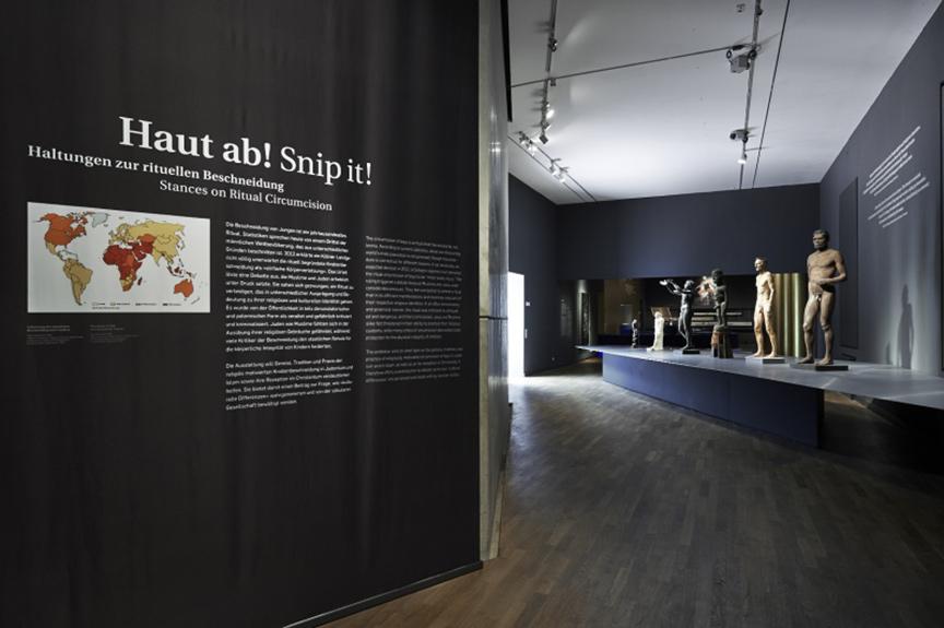 Ausstellungsraum, in dem mehrere nackte männliche Skulpturen auf einem Podest stehen