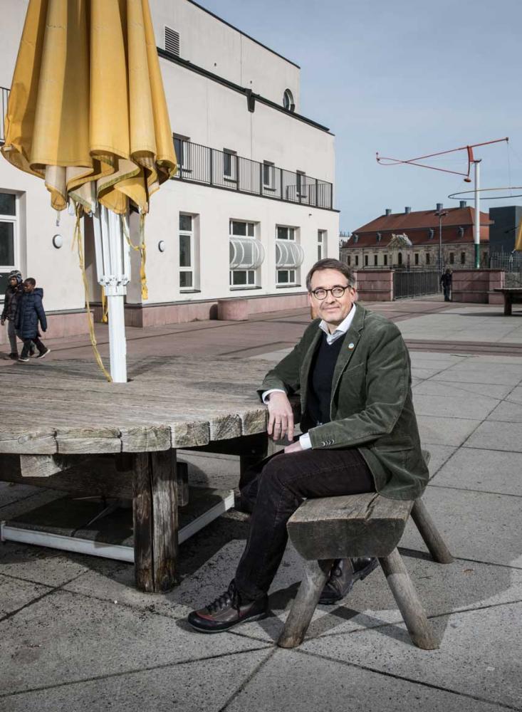 A man with glasses and a green jacket is sitting at a wooden table with a yellow parasol on a roof terrace. The Jewish Museum Berlin can be seen in the background.