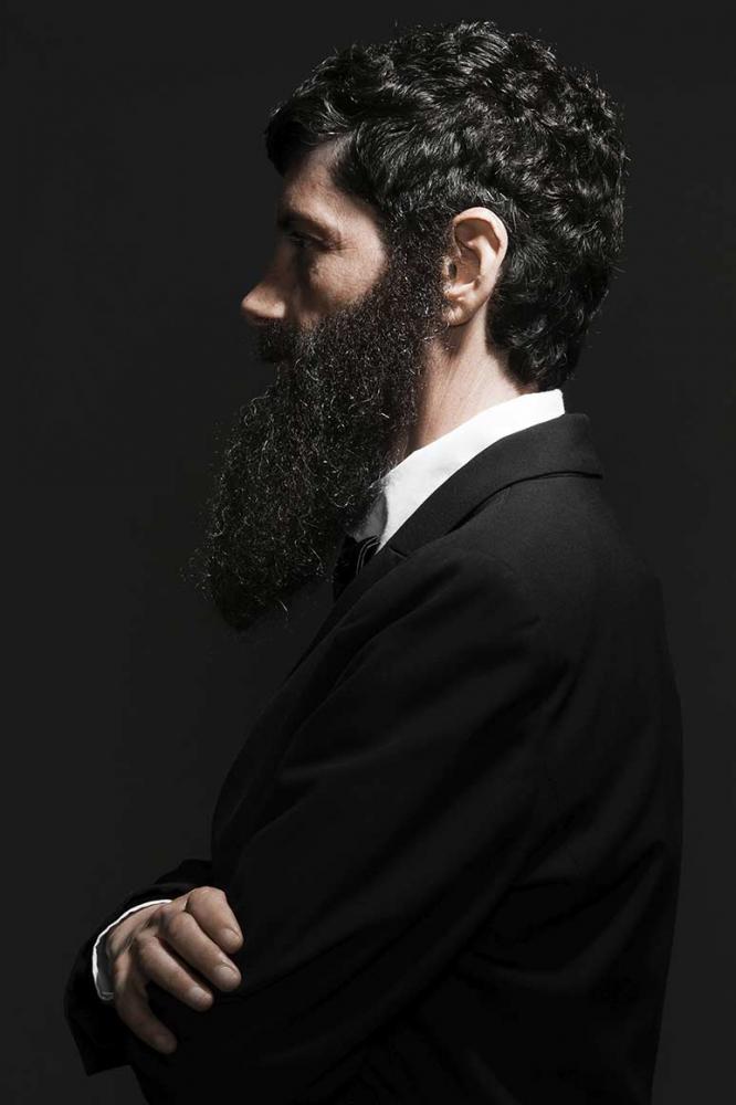 Half-length portrait of a bearded man with long, dark full beard and dark hair in profile. He wears a suit and crosses his arms in front of his chest. The left half of the body is facing the viewer. 