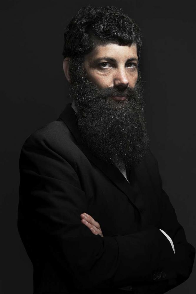 Half-length portrait of a bearded man with long, dark full beard and dark hair. He wears a suit and crosses his arms in front of his chest. The right half of the body is facing the viewer, the face can be seen in half profile.