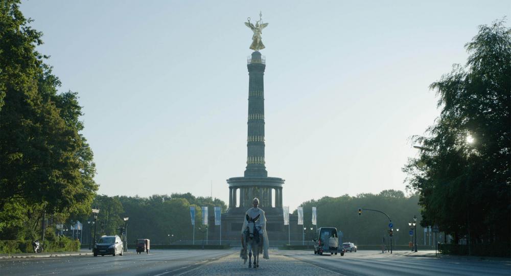 A white-clad, platinum blonde woman rides a donkey on a street. To her right and left are cars, traffic lights and trees of the Berlin Tiergarten. In the background the Berlin Victory Column rises into the sky