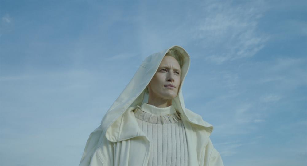 Upper body of an androyn looking person in front of bright blue sky. The cap of her cowl-like robe covers her head