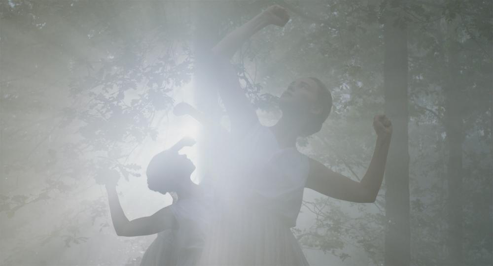 Young women in white sleeveless summer dresses dance in a forest against the light. Their upper arms form a line with their shoulders, the forearms point upwards at right angles with clenched fists