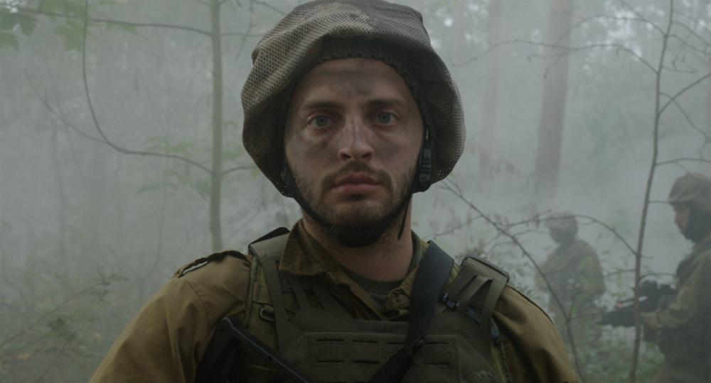 Close-up of head and chest area of a soldier in combat gear in a foggy forest. He wears a helmet on his head and camouflage paint on his face. In the background more heavily armed soldiers can be seen