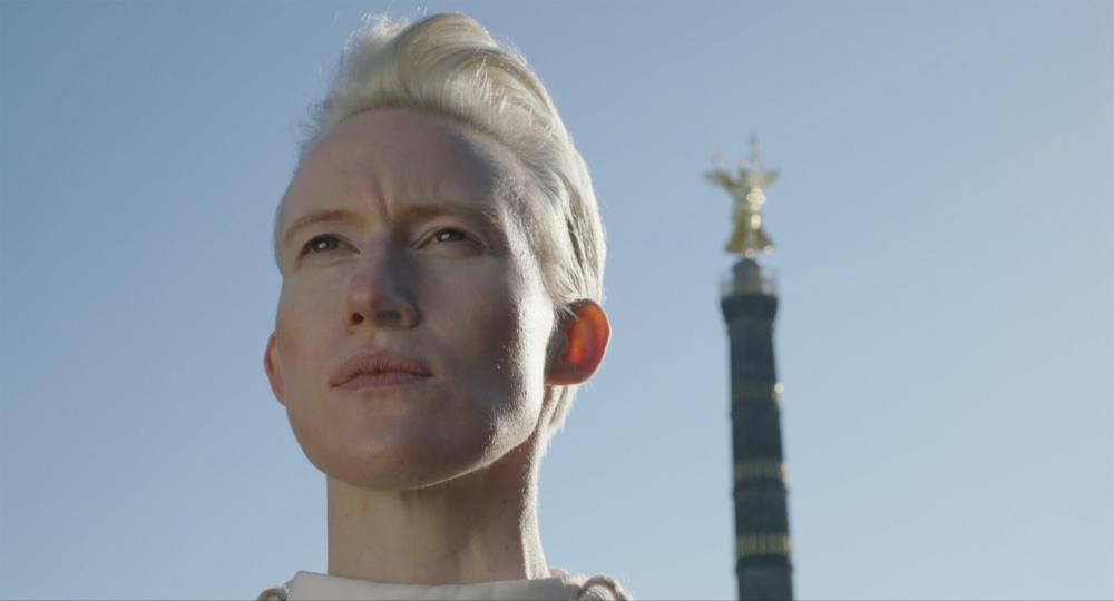 Close-up of the face of a platinum blonde woman with short hair against blue sky. In the background the upper part of the Berlin Victory Column can be seen, but is out of focus