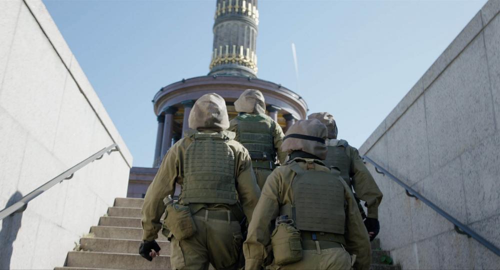 Four people in olive green uniforms with bulletproof vests, helmets and drinking bottles on the stairs of an underpass. You can see the men from behind, the stairs lead up to the Victory Column