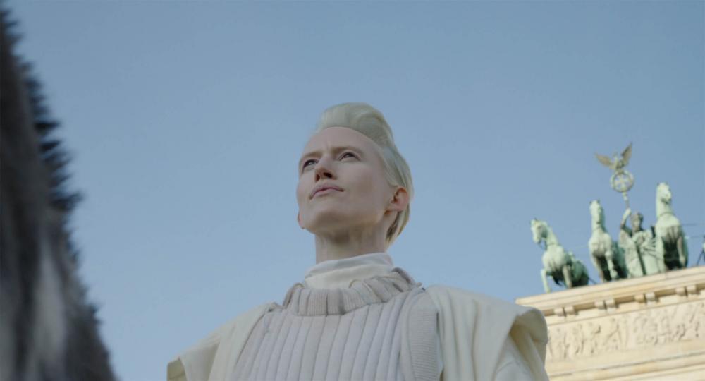 Bust portrait of a platinum blonde woman with short hair and white top in front of bright blue sky. She is photographed from diagonal below, on the right side of the picture you can see the upper part of Berlin Brandenburg Gate