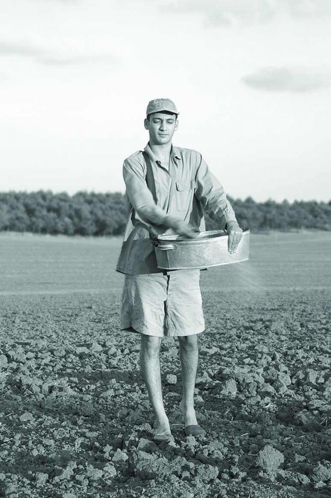 Schwarz-Weiß-Fotografie eines jungen Mannes in kurzen Hosen und Schildmütze. Er steht auf einem frischgepflügten Feld. Aus einer Zinkwanne bringt  er Saatgut aus.