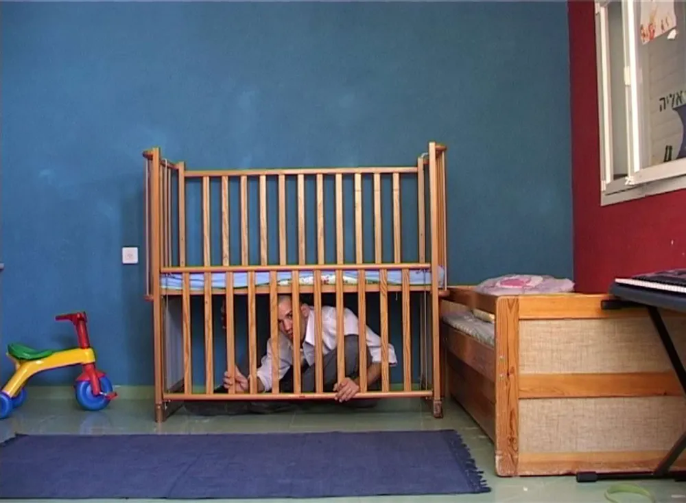 Ein Erwachsener sitzt auf dem Boden eines Kinderzimmers unter dem Gitterbett für Babys und schaut durch die heruntergelassenen Holzgitterstäbe.