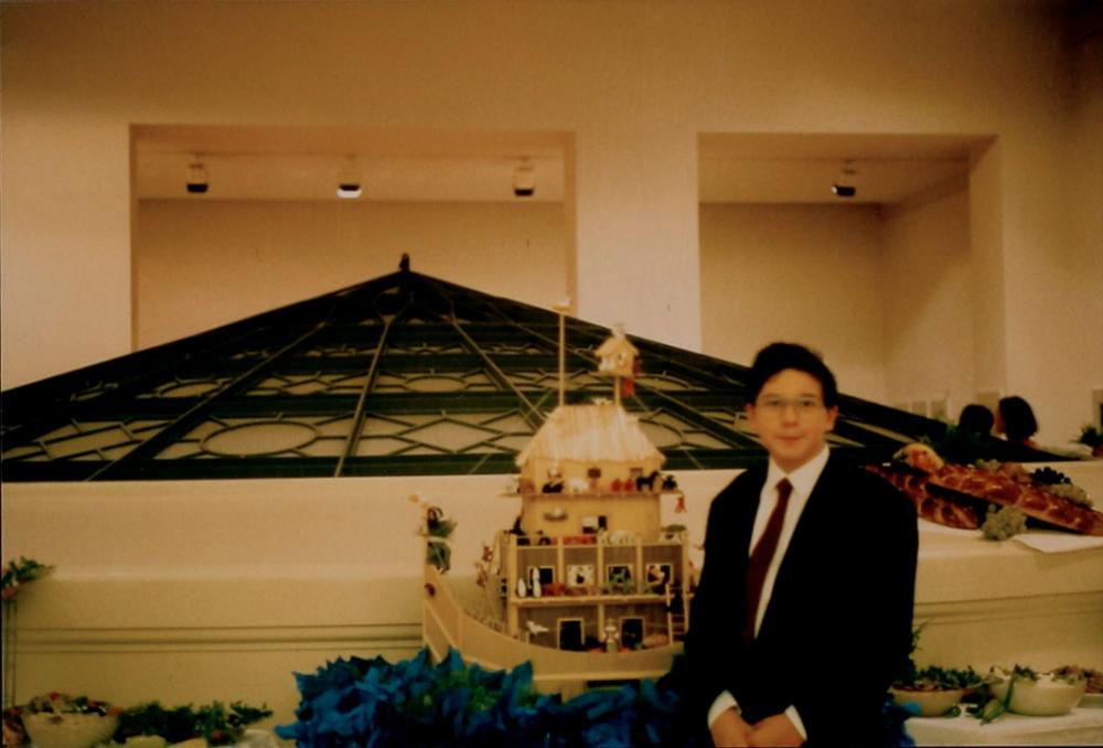 A boy in a dark suit and tie stands next to a toy ark made of wood. A rotunda is visible in the background.