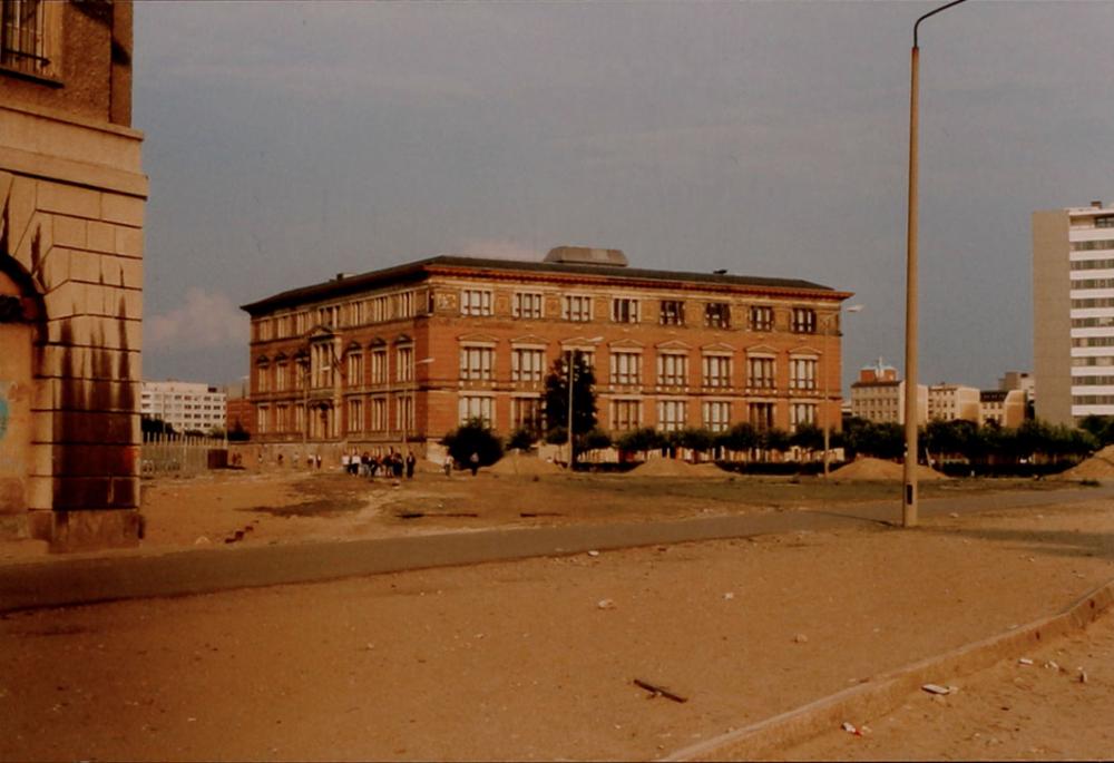 A four-story, square, palace-like structure surrounded by barren land. Apartment buildings are visible in the background. 
