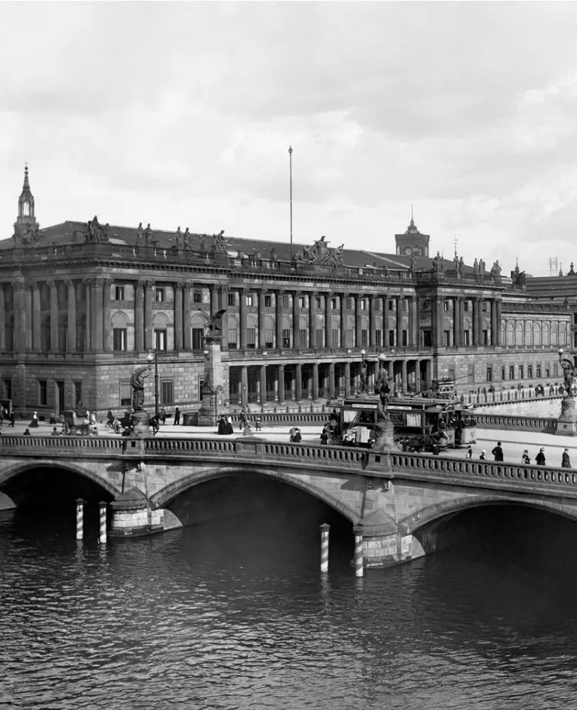 A black and white photograph of a large neoclassical building on a riverbank and a bridge crossing the water.