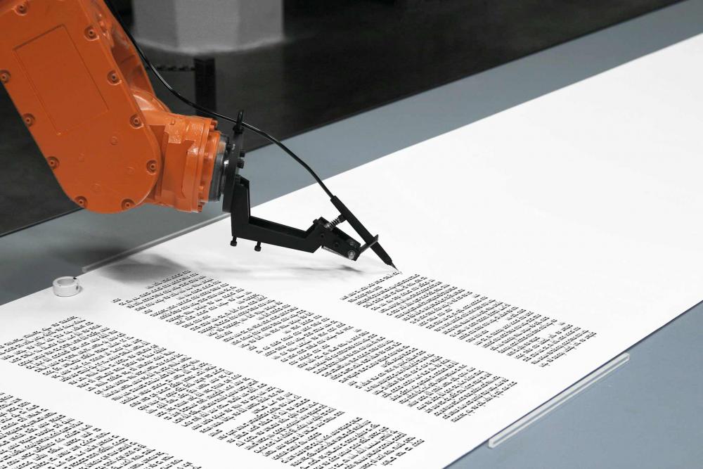 Ein Roboterarm hält eine Tintenfeder und schreibt hebräische Schriftzeichen auf eine lange Papierrolle