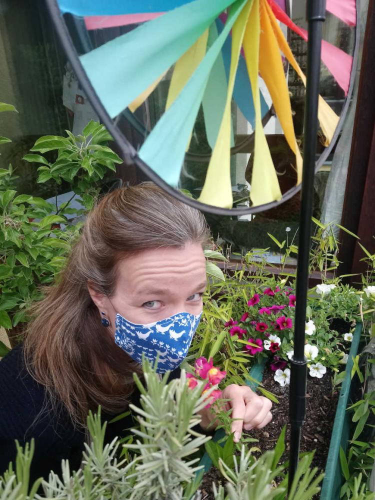 Selfie by Mirjam Bitter with a blue mask, standing on her balcony surrounded by plants - in the foreground a rainbow coloured mill.