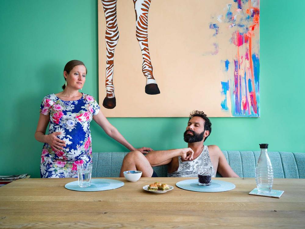 Photography of a pregnant woman in a flower dress and a young man with a dark beard at a dining table, on it three pieces of baklava, on the wall behind them a painting with zebra legs and many colors