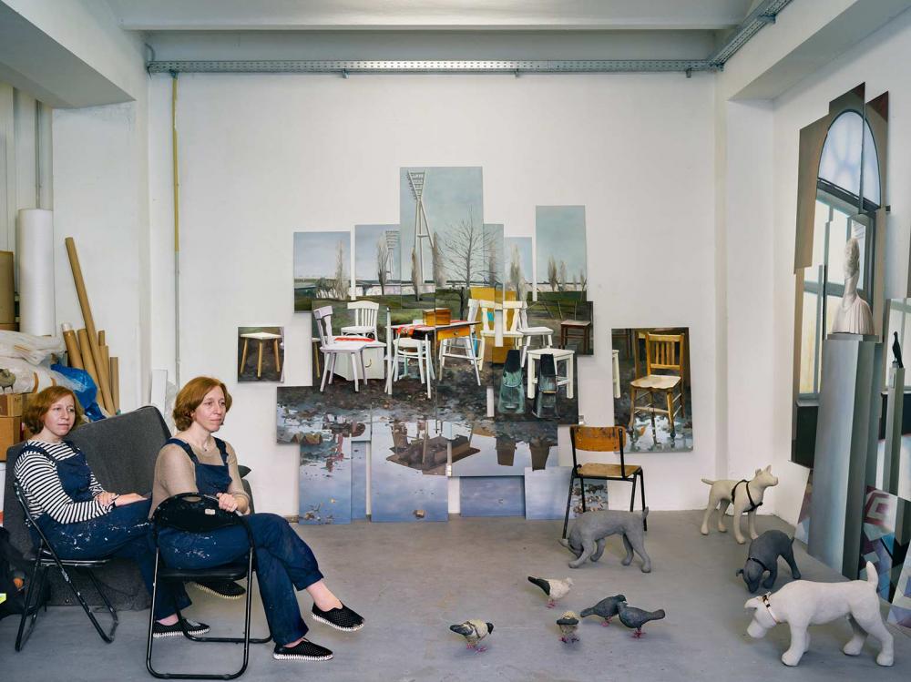 Photography of two twin sisters in blue dungarees on folding chairs in a studio, on the wall a picture with chair motifs, on the floor dog and pigeon sculptures