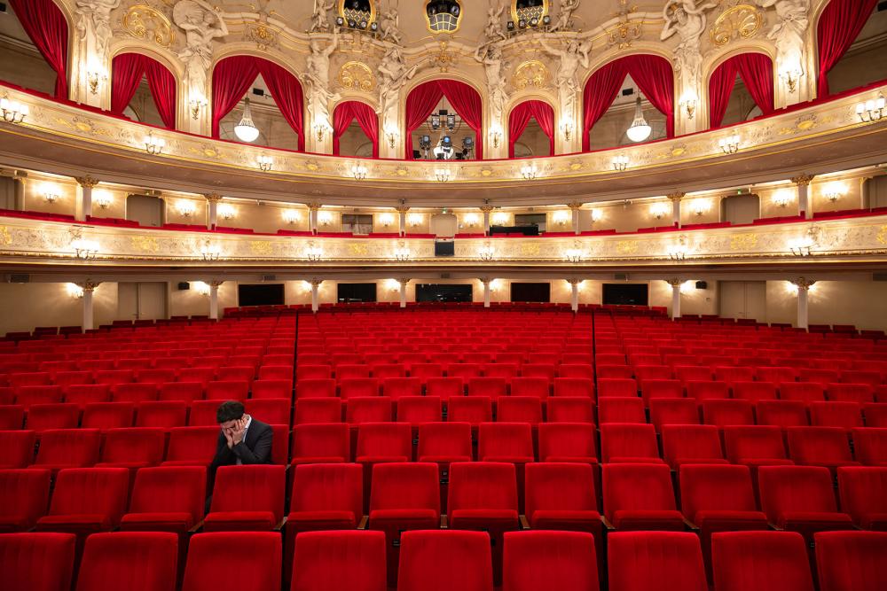 Photography of a person alone on one of the red seats in the audience of an empty theater, resting his head in his hands