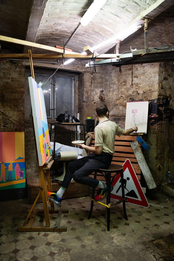 Photography of a person in front of an easel in a basement room, in the background there is a construction site sign on the floor
