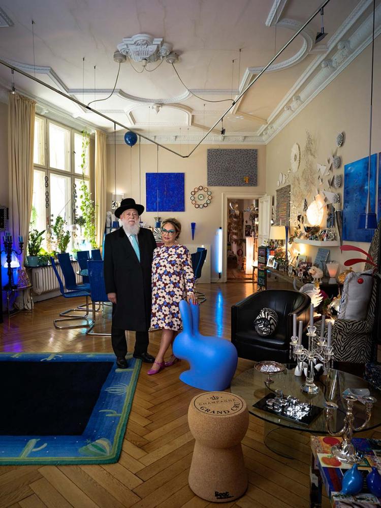 A couple, he in black suit with hat and white beard, she in floral dress with turquoise glasses, pose in a large, very colorfully decorated room, in front of them a blue bunny sculpture