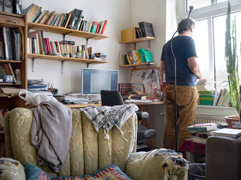 Photo: In a study with bookshelves and computer on the desk, a man stands at the window, his back to the camera