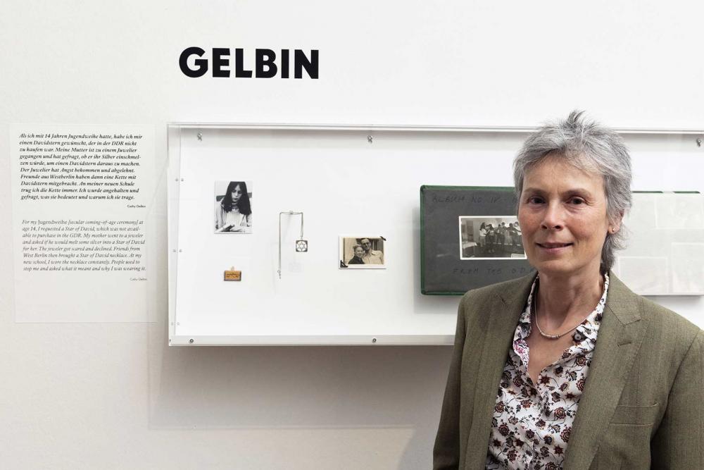 A woman with short gray hair, blouse and jacket in front of a display case with the caption Gelbin and, among other things, a photo of the woman at a young age.
