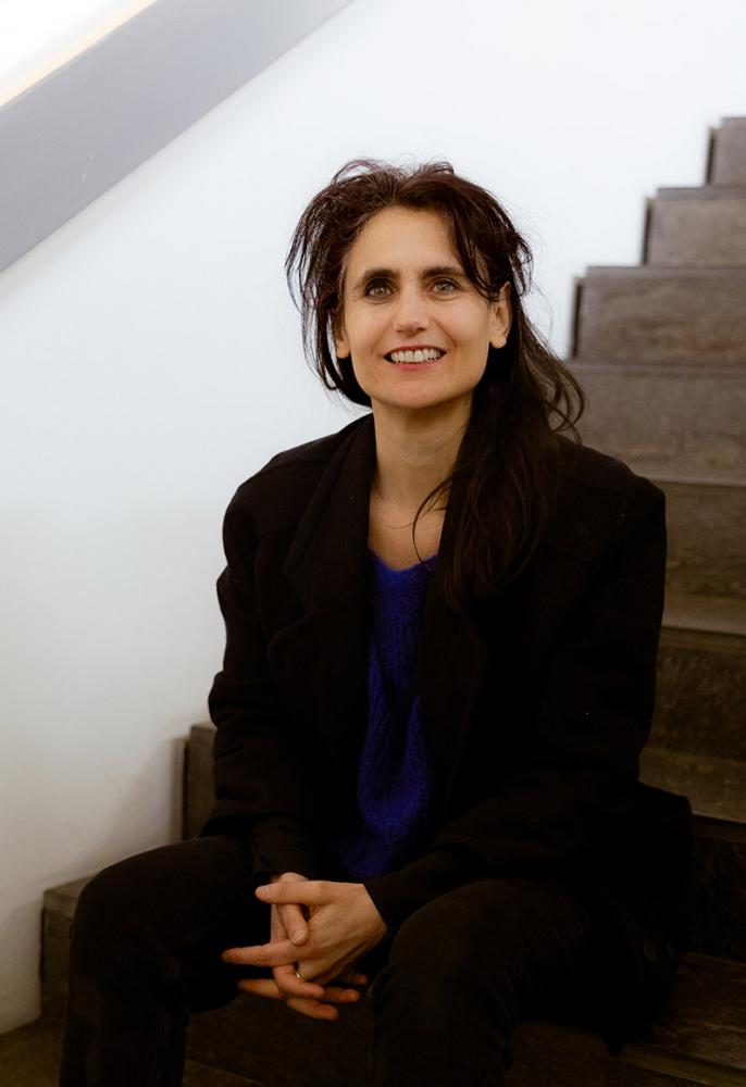 Eine Frau mit langen braunen Haaren, schwarzem Jackett und blauem Pullover sitzt auf einer Treppe und schaut lächelnd direkt in die Kamera.