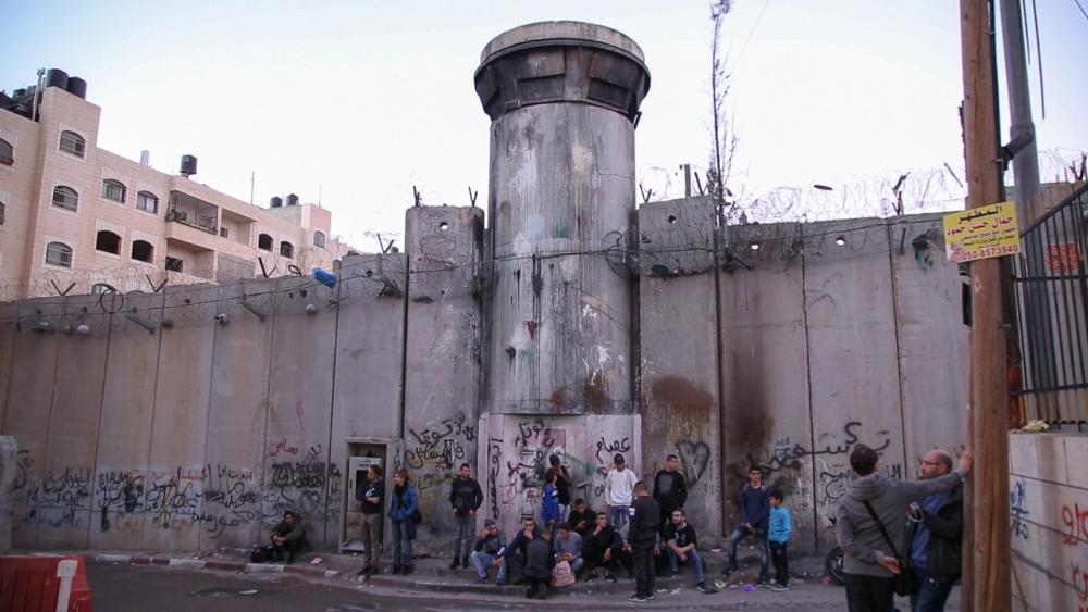 Bedrohlich wirkende Sperrmauer aus Betonfertigteilen mit Wachturm und Stacheldraht, vor der Mauer stehen und sitzen einige Menschen