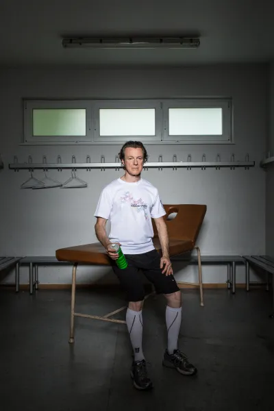 Ein mittelalter Mann in Laufshorts, kniehohen Socken und Trikot blickt in die Kamera. Er sitzt auf einer Liege und h&auml;lt in der rechten Hand eine Wasserflasche. 