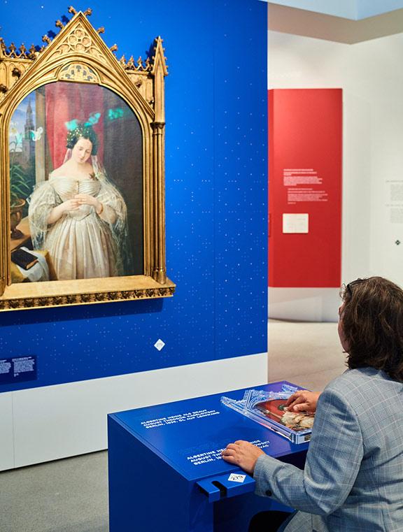 A person runs her fingers over a tactile plan, on the wall in front of her an oil painting of a bride