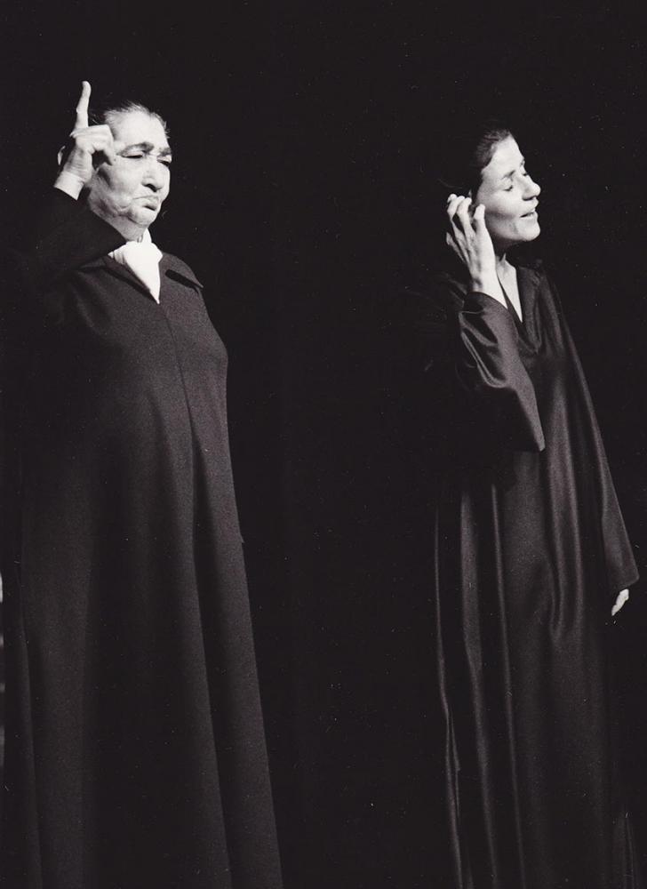 Two women in long black robes stand in front of a black background. One woman raises her index finger, the other brushes her hair behind her ear.